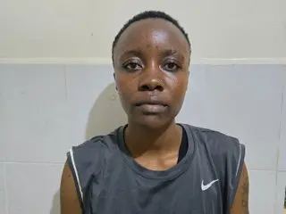 Serious young Black woman in a grey Nike athletic tank top standing against a plain white wall.