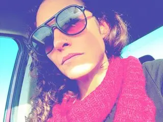Petitemarieethareal poses with her curly hair and sunglasses while wearing a bright pink scarf or top in a car setting.