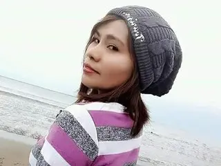PhoebeCream poses with a slight smile, wearing a grey knit beanie and a purple and white striped sweater against a light, neutral background.