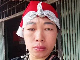 Portrait of a woman wearing a traditional red and white patterned headwrap in a rustic outdoor setting.
