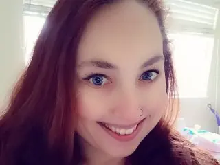 PinkroseBliss smiles closely at the camera, showing her face and long reddish-brown hair in a bright indoor setting.