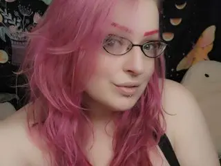 Pinky_Powers poses with her pink hair framing her face, wearing black-rimmed glasses and a black top.