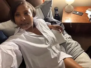 PleasureRebelXXX poses in a white button-down shirt, looking at the camera with her dark hair down in a dimly lit room.