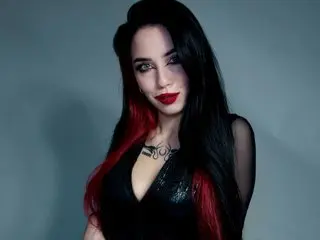 PollyDiva poses with long black hair and red lipstick, wearing a low-cut black top against a plain grey background.