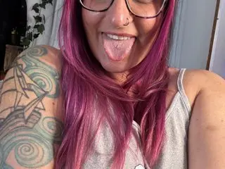 Smiling woman with pink hair and arm tattoo wearing a grey tank top in a bright indoor setting.