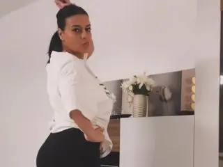 PowerPuff19 poses in a white t-shirt and black bottoms, showing her side profile and dark hair against a plain white wall.
