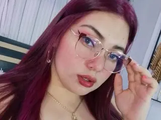 PrettyViolet1 poses close-up, showing her face, red hair, and glasses, wearing a gold necklace against a soft-lit background.