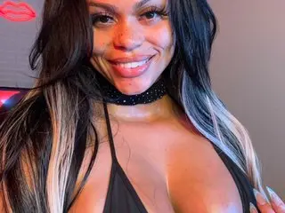 PrincessDalhia smiles while wearing a black choker and a black top, showing her cleavage and long dark hair with blonde highlights.