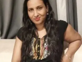 PriyankaBhinde poses with long dark hair, wearing a black sleeveless top with a colorful patterned front, standing against a neutral curtain background.
