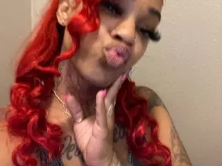 QueeenJ pouts her lips while posing with her hand near her face, showing her long red wavy hair and tattoos.