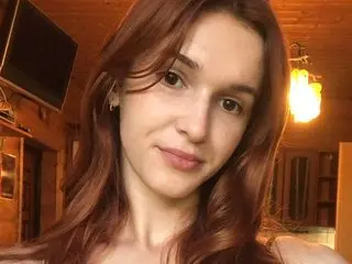 QueenDiiia69 poses with her long red hair down, looking directly at the camera in a warm, indoor setting.