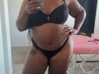 QueenDomi poses in a black bra and matching thong, showing her stomach and pussy, while looking at the camera in a bright room.