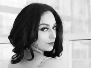 QueenJazminBoheme poses with a side-profile gaze, showing her dark hair and heavy eye makeup against a light, blurred background.