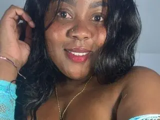 QueenRachelBrown poses close-up, showing her face, dark hair, and bare shoulders or light blue lace top against a plain wall.