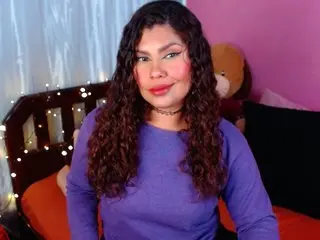 Queenamazon smiles while wearing a purple long-sleeve top with her long, wavy brown hair falling over her shoulders.