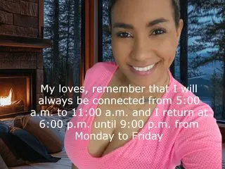 QueenxNefertit smiles while posing in a pink top, showing her face and shoulders against a dark, indoor background.