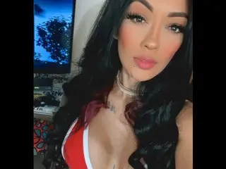 QuinnMPresley poses with long dark hair, wearing a red and white top that shows cleavage, set against a dark indoor background.