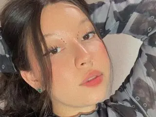 RachelWhiitee poses close-up, showing her face, dark hair, and light freckles against a soft, neutral background.