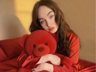 Young woman in a red silk outfit hugging a large red teddy bear on a bed in warm, cozy lighting.