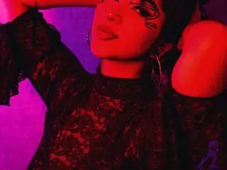 RavenSkys poses with her hands behind her head, wearing a black lace top against a dark background.