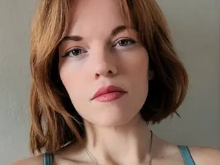RaynaGingerlysnatch poses with her shoulder-length brown hair and red lipstick against a plain wall, wearing a thin teal strap.