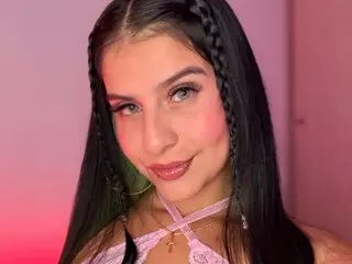 Young woman with long dark hair and braids wearing a pink lace halter top in soft pink lighting.