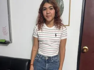 Reimar_ggh poses in a white striped t-shirt and denim shorts, looking at the camera with long wavy hair in an indoor setting.