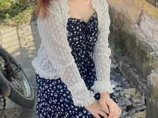 Rena039 poses outdoors wearing a white lace long-sleeve top and a dark floral print dress with her long reddish-brown hair down.