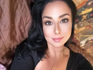 ReneaRaynes poses with a soft smile, showing her long dark hair and cleavage in a black top against a soft brown background.