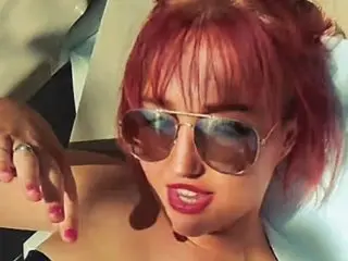 RiaStars poses close-up, showing her red hair and sunglasses while looking directly at the camera with red lipstick.