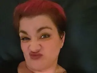 RicanShy pouts her lips and smirks, showing her short red hair and dark eyebrows in a close-up headshot.