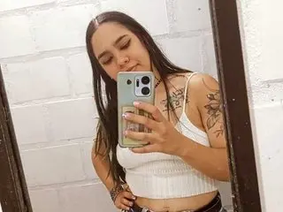 RileySweett poses in a white tank top, showing her long dark hair and arm tattoos against a white brick wall.