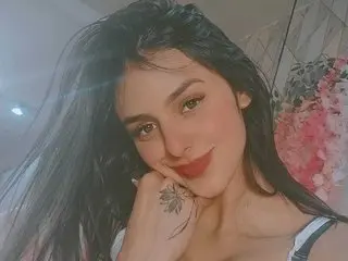 Smiling young woman with long dark hair and a hand tattoo posing in a bright, floral setting.