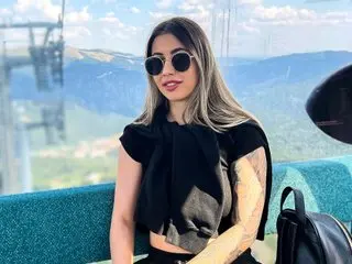 RoseDiamond18 wears sunglasses and a black top while posing outdoors with mountains in the background.