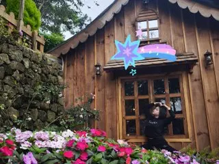 Rose_Mussica stands outside a rustic wooden cabin surrounded by pink and white flowers and green trees.