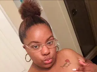 Young Black woman with hair in a high bun and glasses posing indoors with a soft, casual mood.