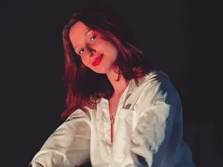 Roussehillss — Roussehillss poses with long dark hair, wearing a white button-down shirt against a dark background with red lighting.