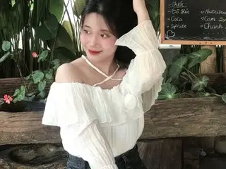 Rove079 poses with her hands near her face, wearing a white off-the-shoulder blouse with long sleeves and dark denim, set against a leafy green background.