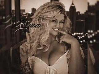 RowanRevnaDisa poses with her hands near her chest, showing cleavage in a white top with her blonde hair styled in waves.
