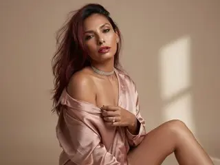 RoxxanneMooree poses in a silky pink robe with her hair down, showing off her cleavage and legs against a neutral background.