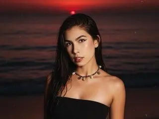 RoxxyReiign poses with long dark hair and a black tube top against a dark sunset beach background.