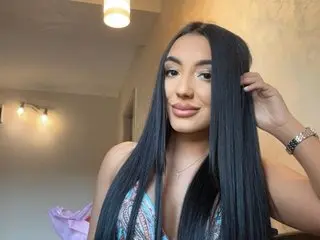 RoxyJensonn with long straight black hair, wearing a patterned bikini top and a silver necklace, posing against a plain wall.