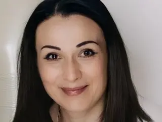 RozmarinnBeauty smiling at the camera with long dark hair and a neutral background.