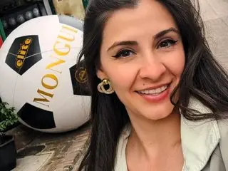 Smiling brunette woman in a white jacket posing next to a large Mongui branded soccer ball outdoors.