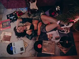 RubiKayale lies on a bed wearing a black bra and shorts, showing her cleavage and legs while lying next to vinyl records.
