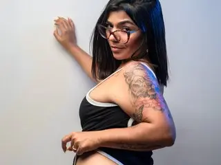 RubyMoonn poses against a white wall, wearing a black bodysuit that shows off her shoulders and cleavage, with her dark hair styled down.