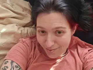 Rubyred48 smiles for a close-up selfie while laying on tan bedding, wearing a pink t-shirt and showing her dark hair.