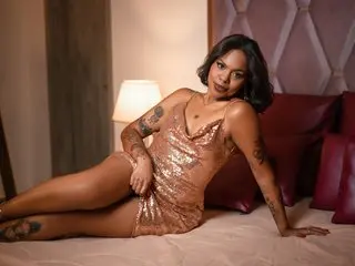 RutFletcher poses seductively in a bronze shiny slip dress, showing off her tanned skin and dark hair while sitting on a bed.