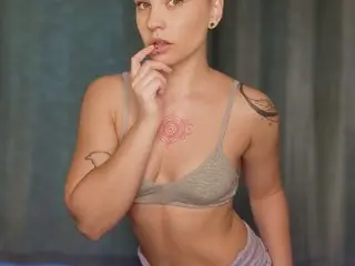 RylieParker poses with her hand near her mouth, wearing a grey bra and showing off her tattoos against a dark background.