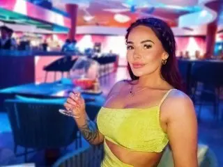 SOAKED_GYMGIRL_MILF_UK is a brunette woman in a yellow crop top sitting at a bar with a drink in her hand.